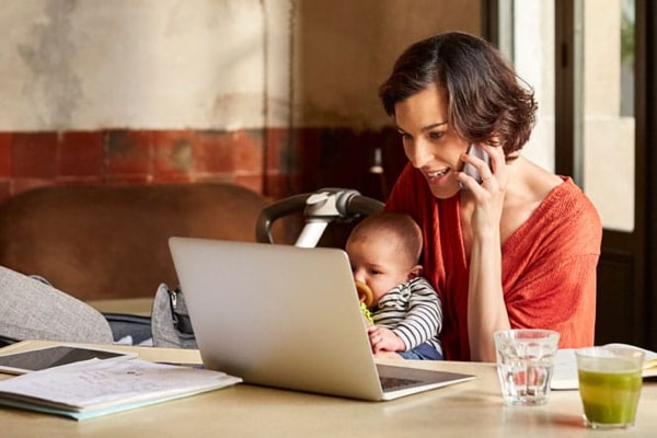 An adult holding a baby, looking at a laptop screen, and talking on the phone