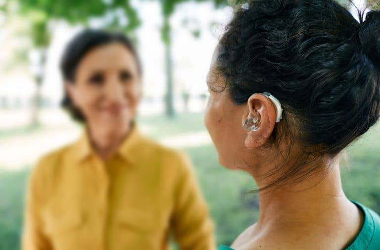 Adult woman with a hearing impairment uses a hearing aid to communicate with her female friend at city park. Hearing solutions
