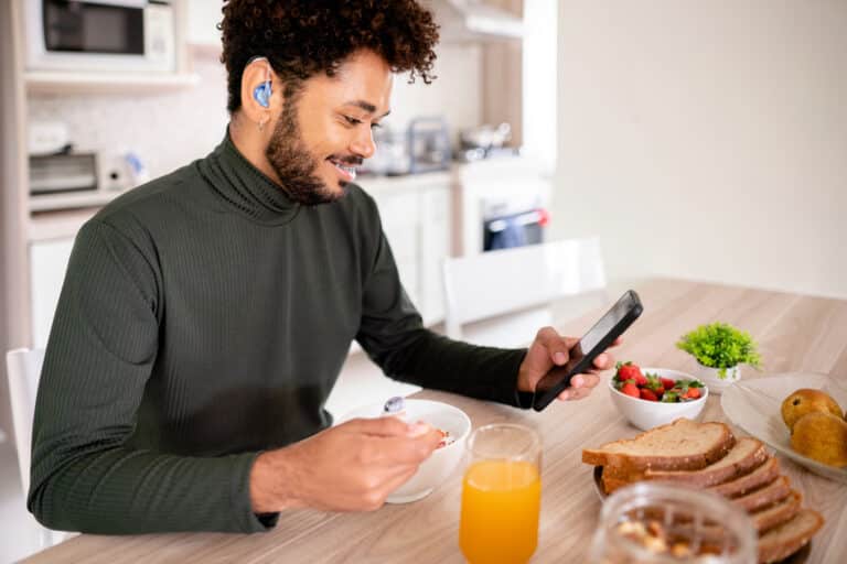 Young man with a hearing aid looks at his smartphone over breakfast.