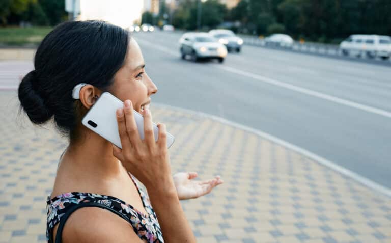 Adult woman with a hearing impairment uses a hearing aid in everyday life, talking on phone in urban city outdoor.