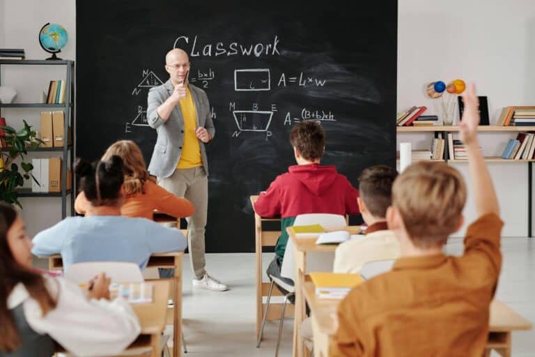 Teacher in front of the classroom calling on a student with their hand up.